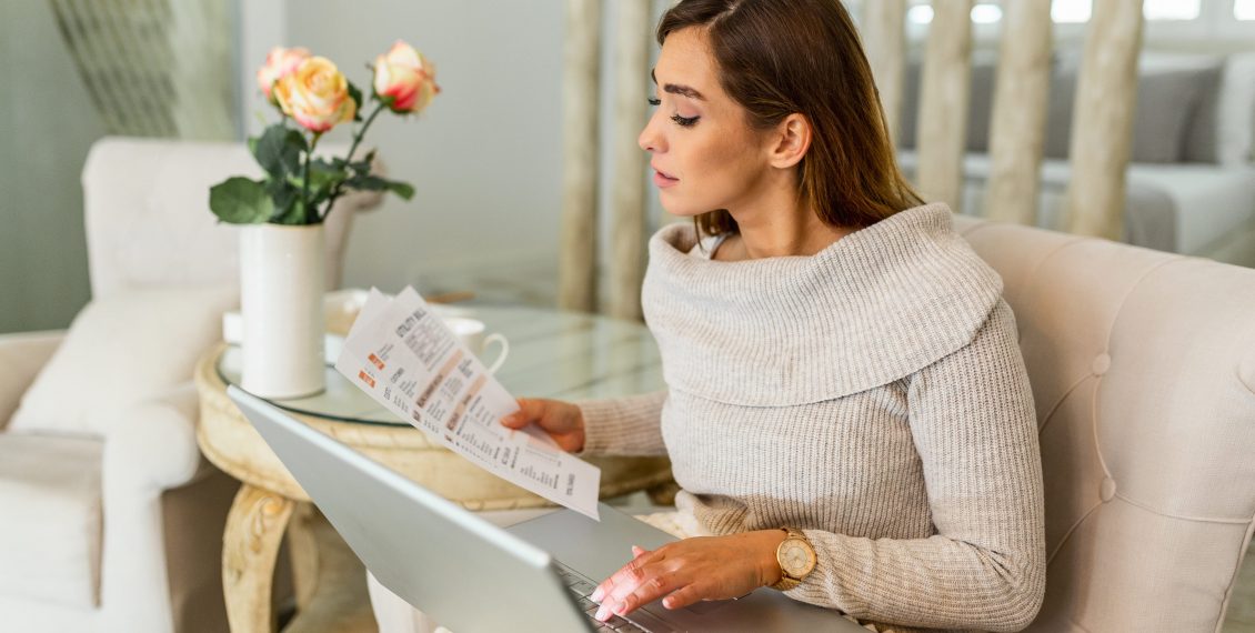 Casually dressed young woman holding papers in her hands, calculating and paying bills at home. Happy beautiful woman sitting on the sofa with laptop calculating home finances. Work from home
